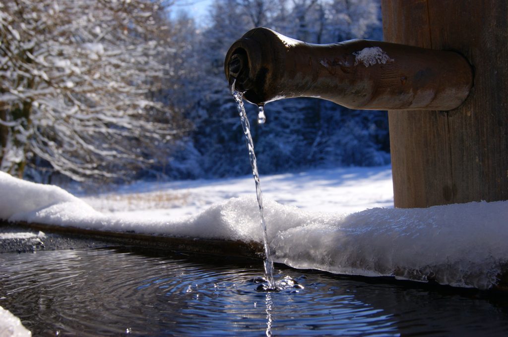Aus einem Brunneneinlauf fließt Wasser in ein Auffangbecken vor einer winterlichen Landschaft.