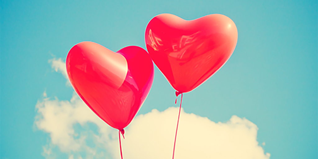 Zwei rote Herzluftballons, die mit Gas gefüllt sind, schweben an roten Schnüren vor blauem Himmel.