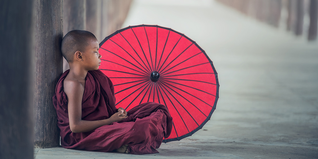 Ein Junge in asiatischer Mönchskleidung sitzt an einen Holzpfahl gelehnt neben einem roten Schirm.